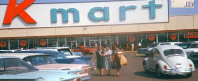 A vintage scene of a Kmart store with a large "Kmart" sign. Shoppers walk in the parking lot among classic cars, including a Volkswagen Beetle. A price tag in the corner shows 88 cents in retro styling.