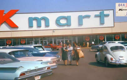 A vintage scene of a Kmart store with a large "Kmart" sign. Shoppers walk in the parking lot among classic cars, including a Volkswagen Beetle. A price tag in the corner shows 88 cents in retro styling.