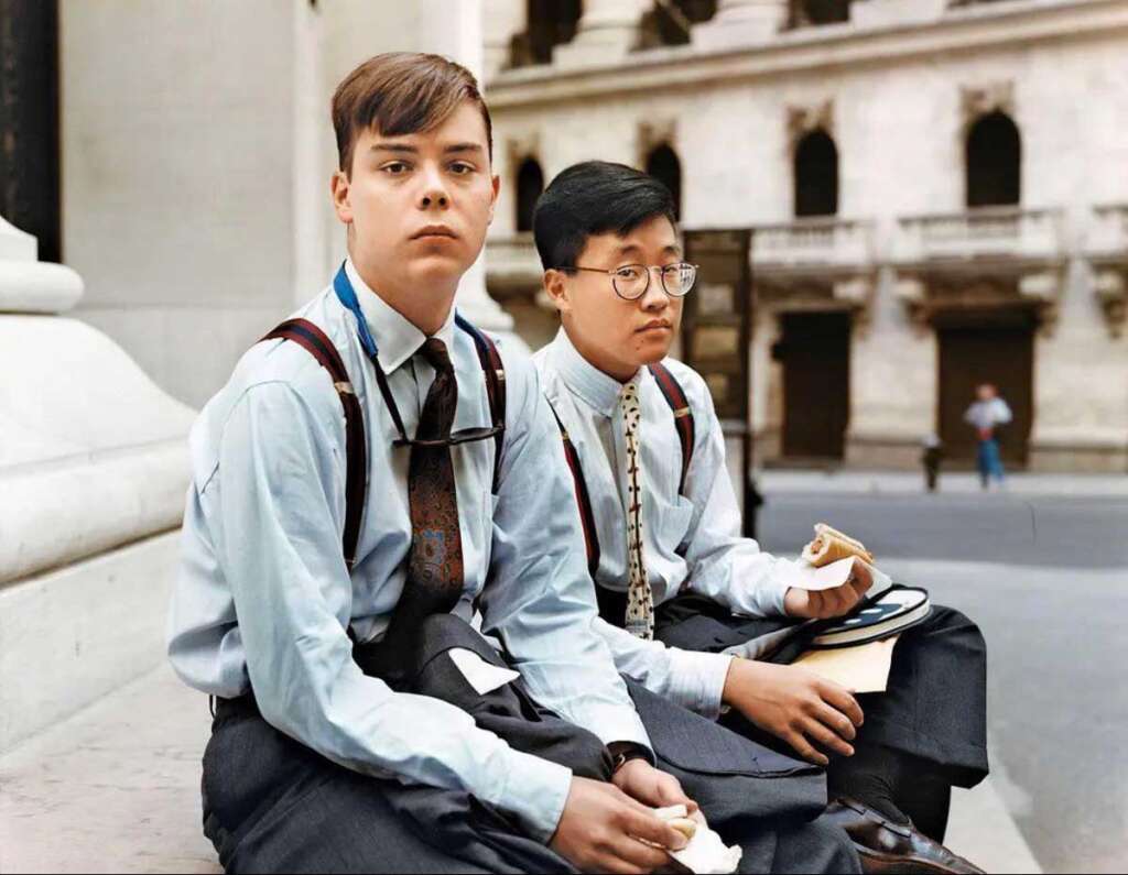 Two young men in formal attire sit on steps, eating sandwiches. One has short brown hair and suspenders, the other has glasses and a tie with a white shirt. They are in an urban setting with classical architecture in the background.