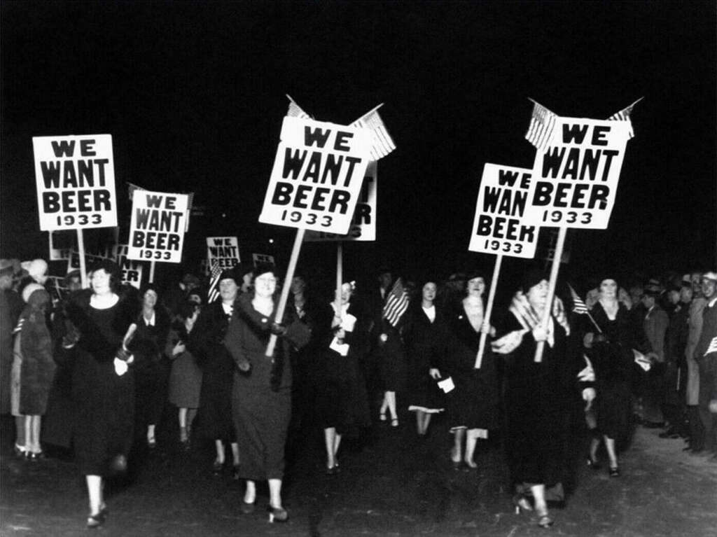 Black and white photo of a 1933 protest. A group of people marches, holding signs that read, "WE WANT BEER." The crowd mostly wears dark clothing, and several American flags are visible.