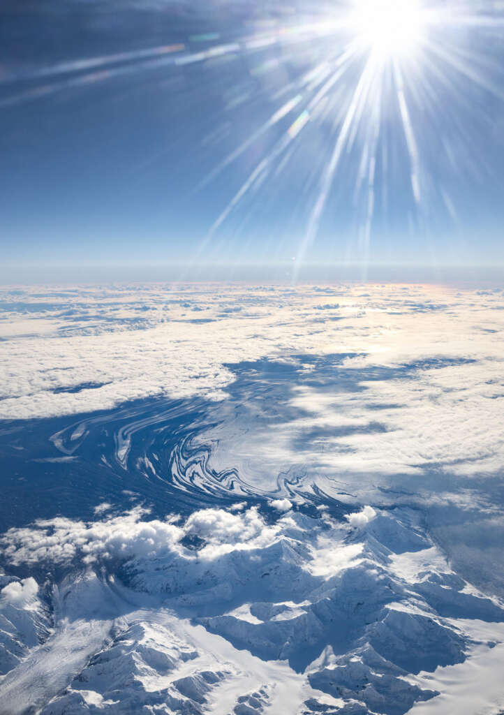 Aerial view of a vast icy landscape with swirling patterns on the surface, snow-covered mountains in the foreground, and a bright sun shining overhead, illuminating the sky with sun rays and casting shadows across the scene.