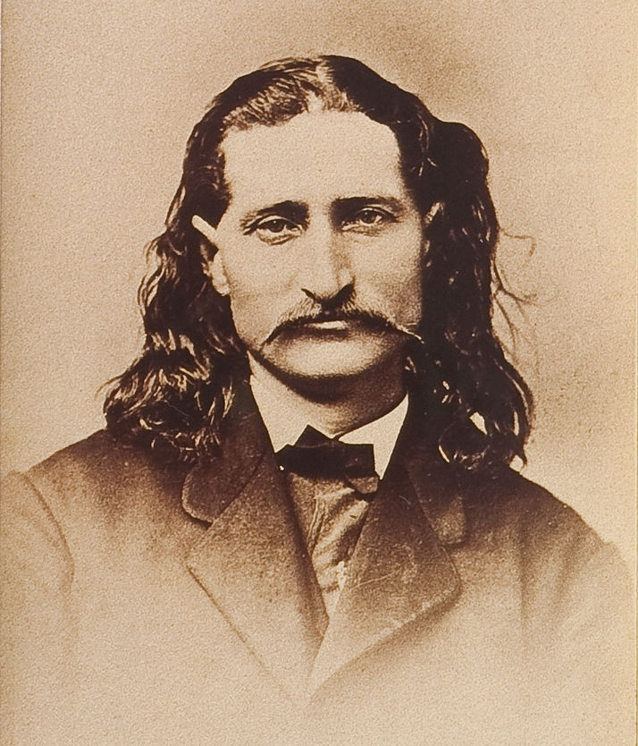 A sepia-toned vintage portrait of a man with long hair and a mustache, dressed in a suit with a bow tie, looking directly at the camera against a plain background.