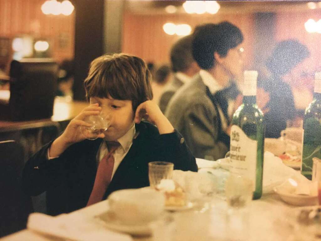 A child in a formal suit drinks from a glass at a dining table with bottles, dishes, and glasses. Other people are seated in the background. The setting appears to be a restaurant with dim lighting.