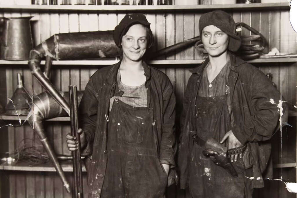 Two women wearing work overalls and caps stand in front of shelves lined with industrial tools and equipment. One holds a long cylindrical object, while the other rests her hand on her hip. Both wear dust-covered clothes and smile at the camera.