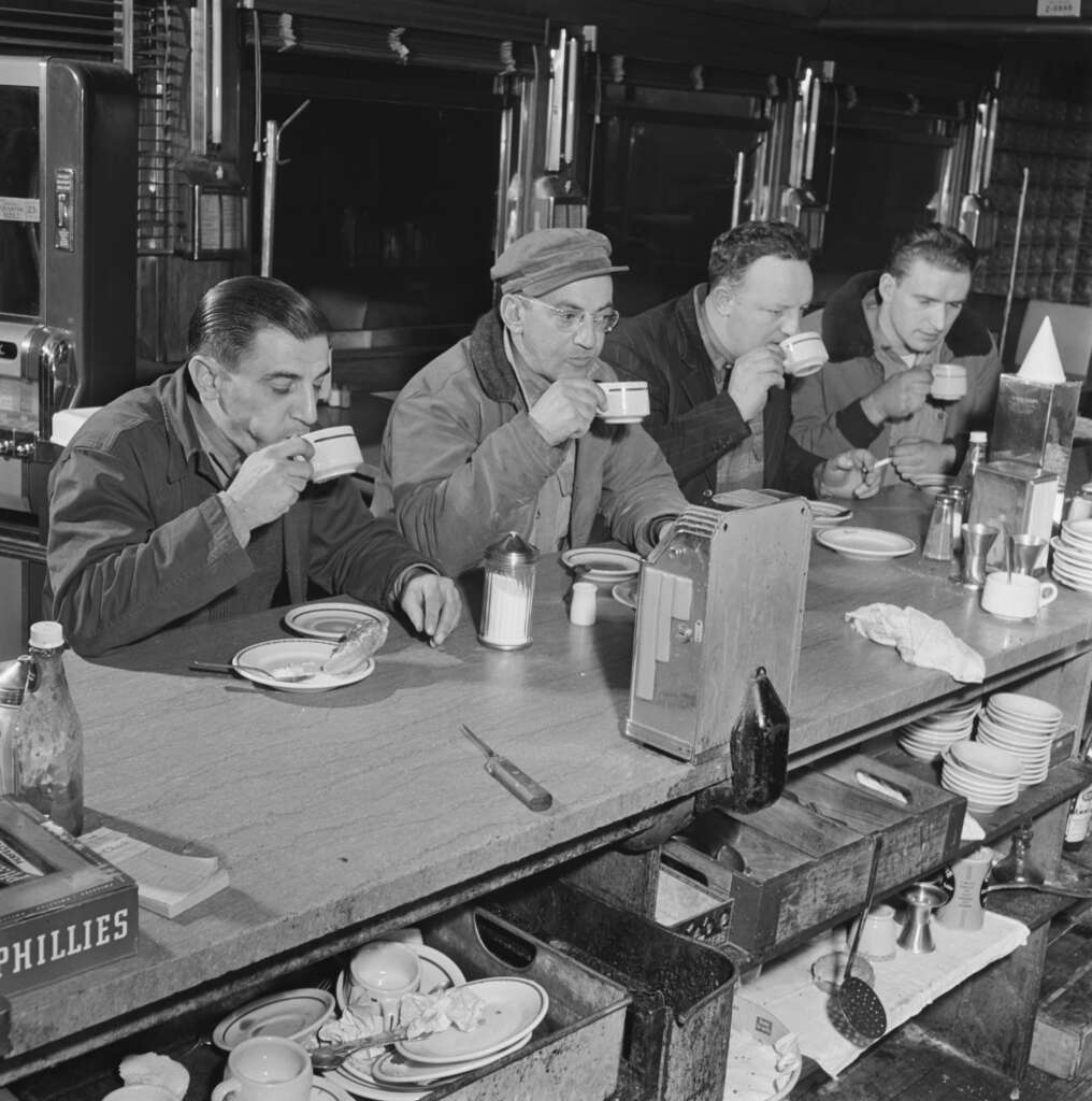 Four men in work attire sit at a diner counter, each sipping from a mug. The counter has plates, condiments, a napkin holder, and a jukebox. The men appear to be taking a break, engaged in relaxed conversation.