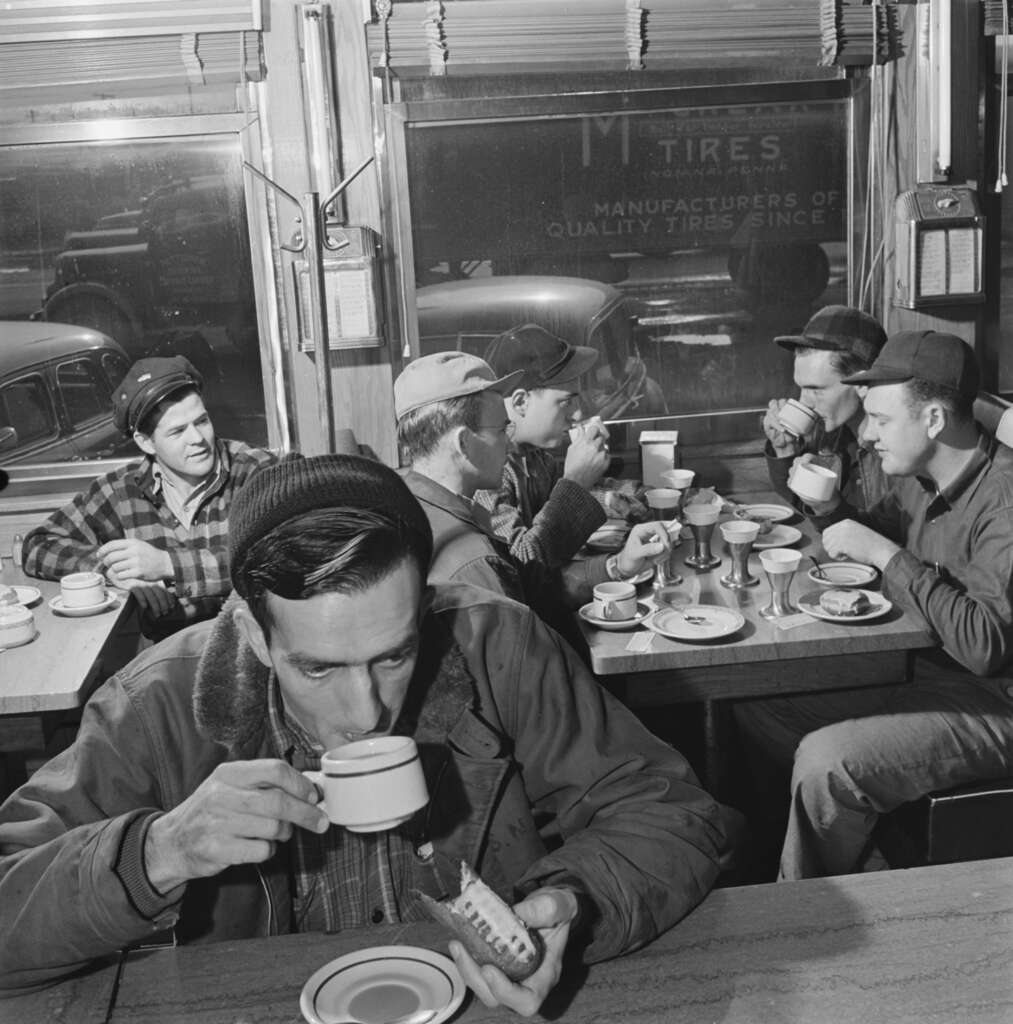 A group of men in casual jackets and hats sit at a diner counter, drinking coffee and holding food. The diner has a window with a sign advertising quality tires, vintage cars visible outside. The scene is in black and white.