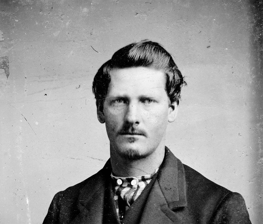 A black and white portrait of a man with short hair and a mustache, wearing a dark suit and patterned tie. The background is plain, and the photo has a vintage, slightly worn appearance.