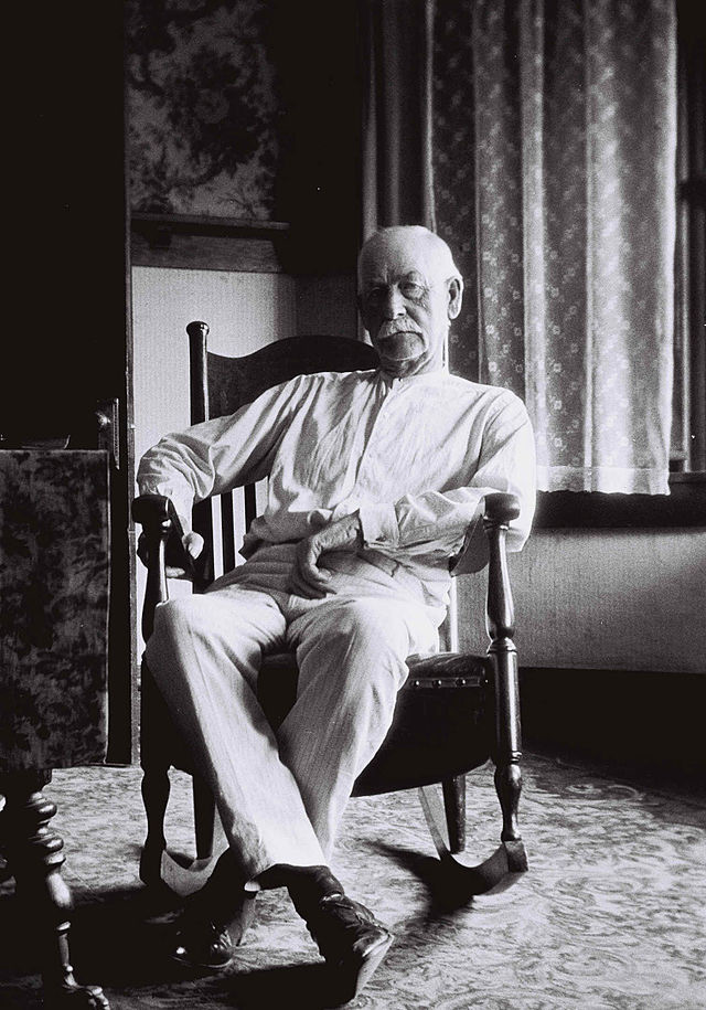 An elderly man with a bald head and mustache sits in a wooden rocking chair, wearing a light-colored shirt and trousers. He is indoors, with patterned wallpaper and a curtain visible behind him. The photo is in black and white.