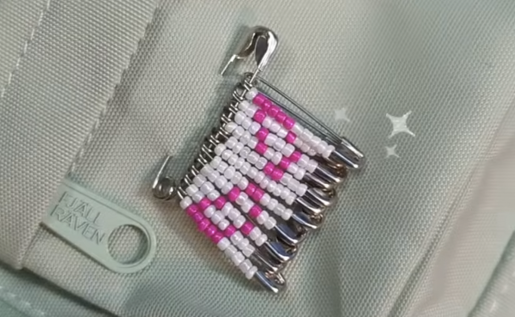 A series of safety pins attached to a green fabric, forming a design with white and pink beads. There are faint star designs near the pins and a Fjällräven tag nearby.