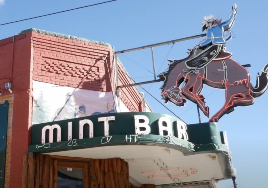 Sign for "Mint Bar" shows a neon cowboy on a bucking horse above the entrance of a brick building with a green roof trim. The sign is outlined against a clear blue sky.