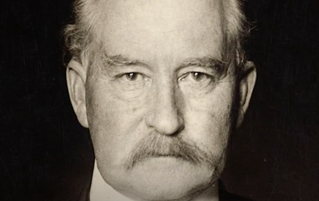 Black and white photograph of an older man with a serious expression, wearing a suit and tie. He has a thick mustache and short hair. The background is dark, focusing attention on his face.