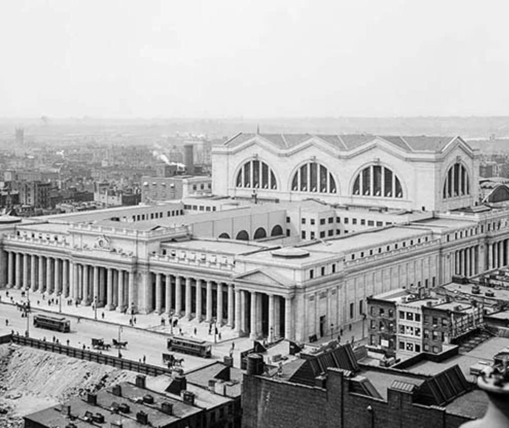 A historical black-and-white aerial photograph of a large neoclassical building with columns and arches, surrounded by early 20th-century city structures and streets, with people and horse-drawn carriages visible in the foreground.