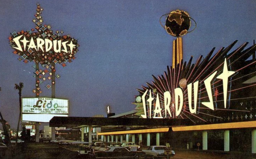 Vintage photo of the Stardust Hotel and Casino in Las Vegas at dusk. Neon signs glow, with the word "Stardust" prominently displayed. Cars are parked in front, and a globe adorns the building's rooftop.