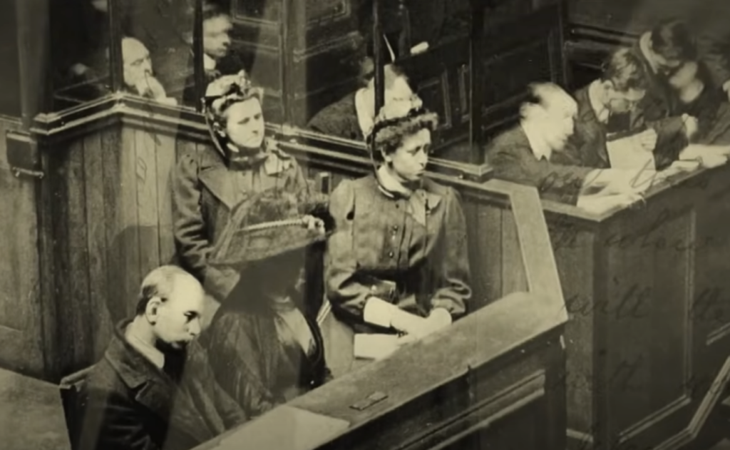 A vintage courtroom scene with four women sitting in a jury box wearing dark historical clothing and hats. Two men are seated in front of them, one writing on a notepad. The background features wooden paneling.