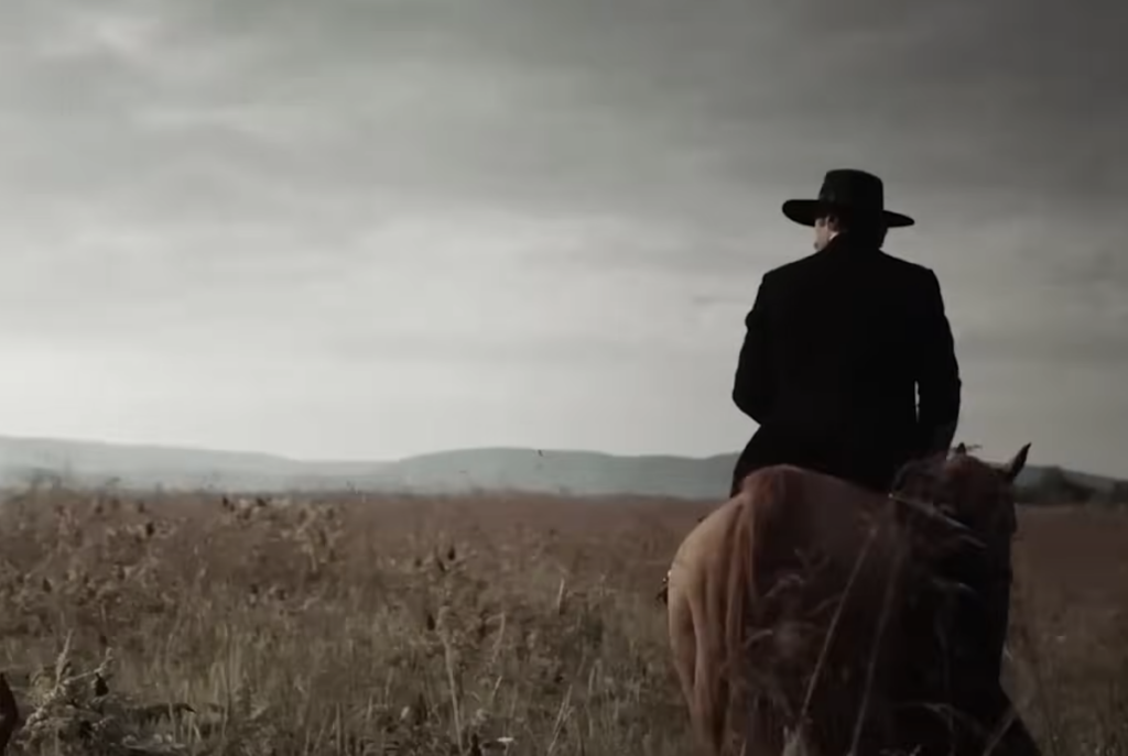 A person in a black hat and coat rides a brown horse through a grassy field under a cloudy sky. The landscape is expansive, with distant hills visible on the horizon. The scene conveys a sense of solitude and vastness.