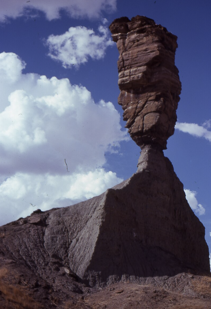 Tall rock formation resembling a teetering tower against a cloudy sky. The base is narrow, widening out towards a large, flat top. The structure appears precariously balanced on a sloped rocky pedestal.