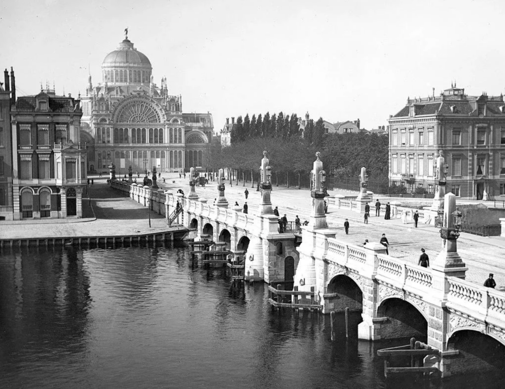 Black and white photo of an ornate stone bridge with decorative columns, spanning a river. In the background, a grand domed building and symmetrical architecture are visible, surrounded by trees and people walking along the paths.