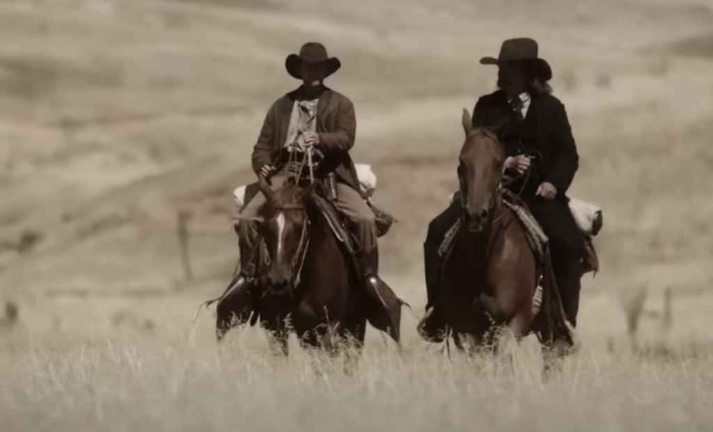 Two cowboys on horseback ride through a grassy, golden plain. They wear wide-brimmed hats and rustic clothing, suggesting a historical or western setting. The landscape is vast and open under a clear sky.