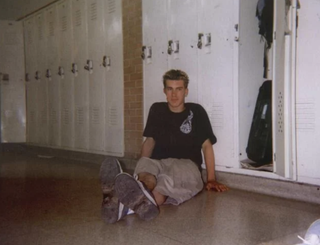 A person sits on the floor in a hallway lined with white lockers. They wear a black t-shirt and beige shorts, and their feet are stretched out. An open locker with a backpack inside is visible in the background.