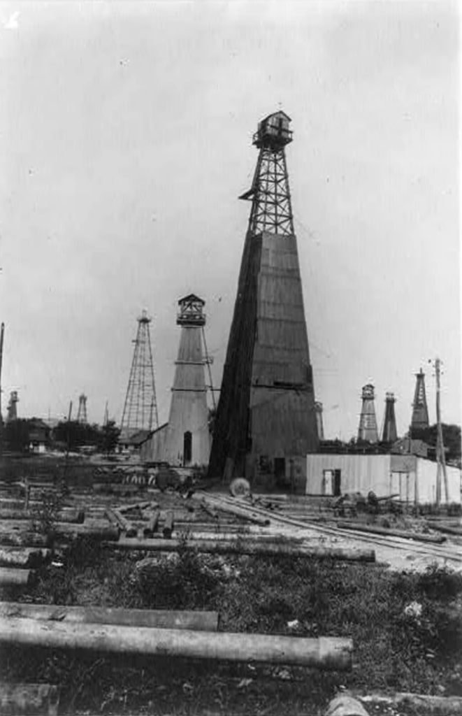 Black and white photo of an oil field with multiple wooden oil derricks. The derricks are surrounded by scattered logs and wooden structures. The sky is overcast, and the landscape appears flat and industrial.