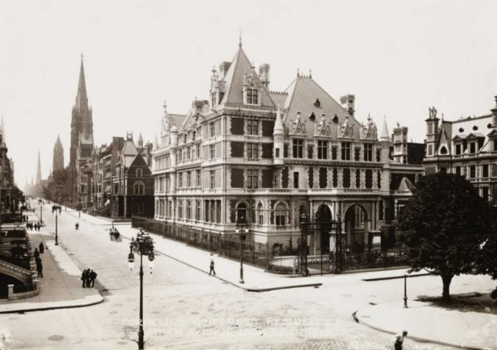 A historic black and white photograph depicting a grand, ornate building on a corner with multiple spires and arches. The street is cobblestone with a few pedestrians and lamp posts. There are several other gothic-style buildings in the background.