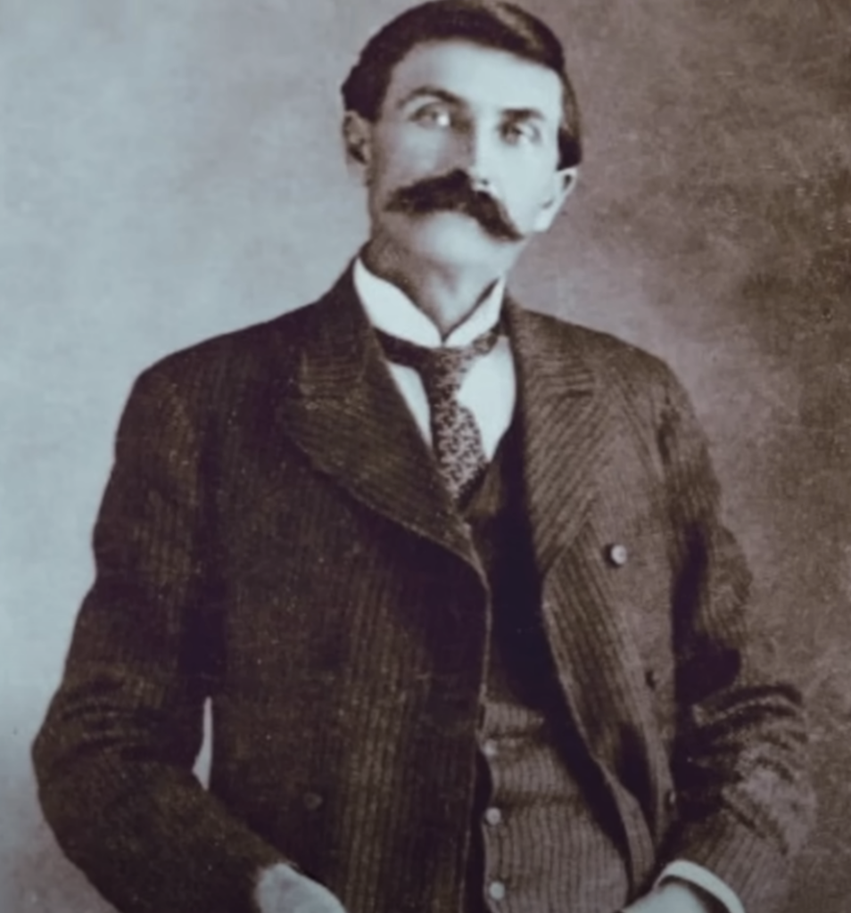 Vintage photograph of a man with a mustache, wearing a dark suit, vest, and tie. He stands with one hand in his pocket, gazing slightly to the side. The image has a sepia tone, indicating its historical age.