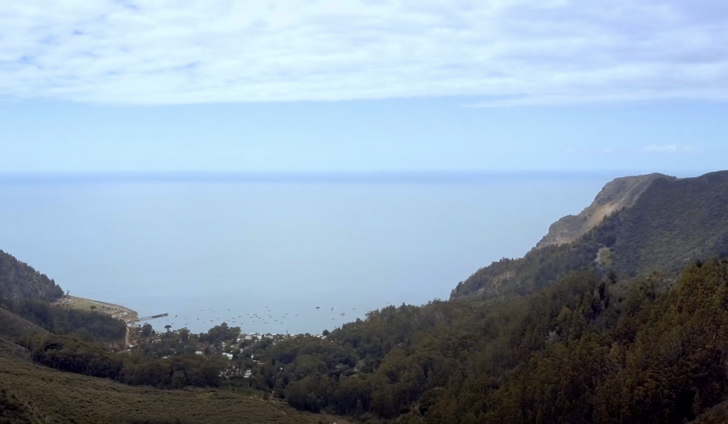 Aerial view of a coastal landscape with a calm sea horizon. Forested hills frame the scene, and a small town with a marina is visible near the shore. The sky is partly cloudy, creating a serene and expansive atmosphere.