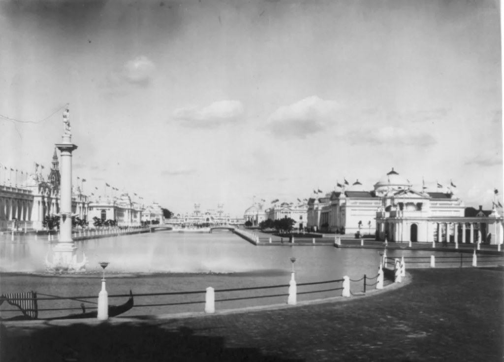 Black and white photo of the Panama–Pacific International Exposition grounds, showing a large reflecting pool surrounded by ornate buildings with domes and columns. Flags adorn the structures, and fluffy clouds dot the clear sky.