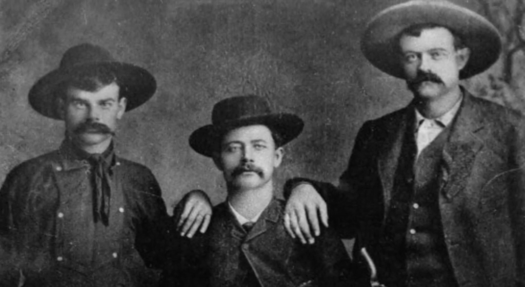 Three men from the late 19th or early 20th century pose for a black-and-white portrait. They wear hats and suits typical of the era. The man in the middle is seated, while the other two stand on either side, each with a hand on his shoulder.