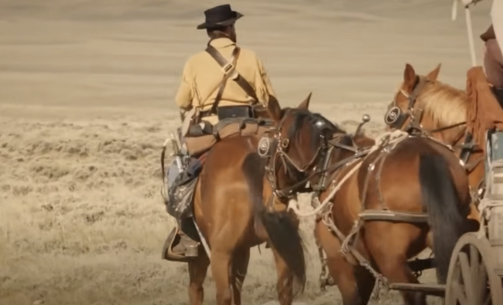 A person in a yellow shirt and black hat rides a brown horse, accompanied by another person on horseback and a third horse. They are traveling through a dry, open landscape with a covered wagon to the right.