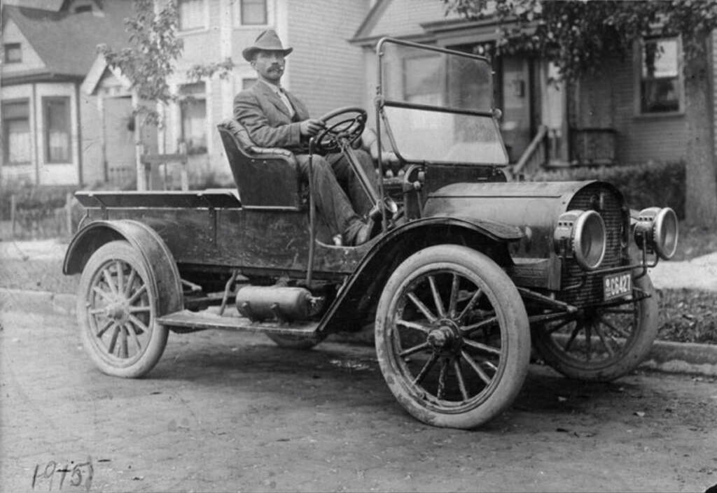 A vintage black and white photograph of a man sitting in an early 20th-century open-top car. The car has large spoked wheels and is parked on a residential street. Houses and trees are visible in the background.