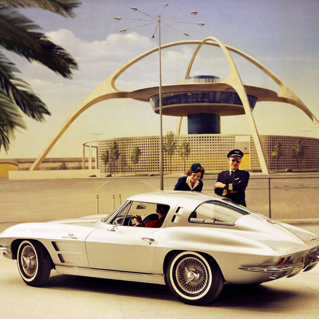 A classic silver sports car is parked in front of a futuristic, arched building. Two people in airline uniforms, a woman and a man, stand next to it, appearing engaged in conversation. The sky is clear, and palm leaves are visible in the foreground.