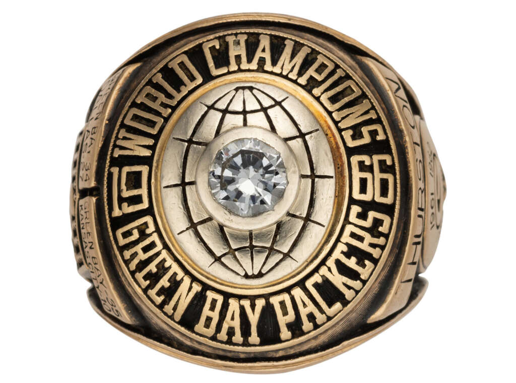 Close-up of a gold Green Bay Packers championship ring with a large central diamond. The ring is inscribed with "World Champions 66 67" and shows the team's name prominently.