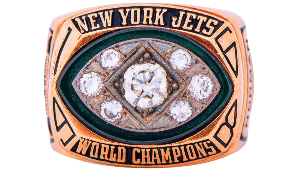 Gold championship ring with "New York Jets" and "World Champions" engraved. Features a large central diamond surrounded by smaller diamonds on a green background, shaped like a football field.