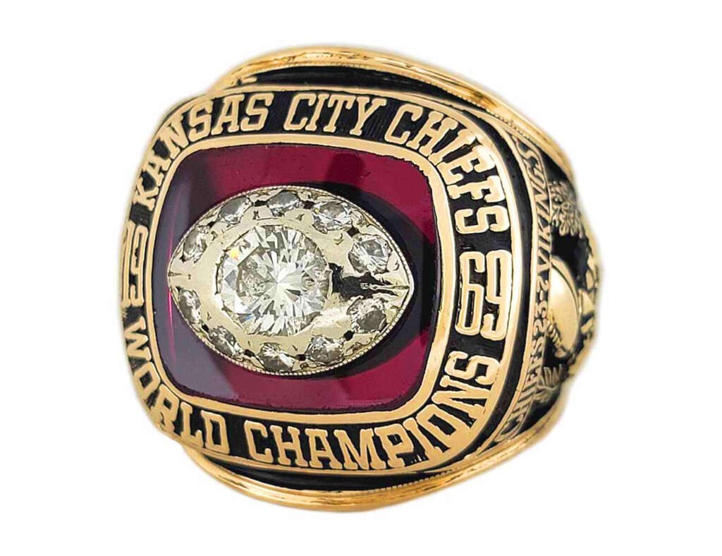 A gold Kansas City Chiefs championship ring with a large central diamond and several surrounding diamonds set in a red background. The ring is engraved with "KANSAS CITY CHIEFS", "WORLD CHAMPIONS", and the numbers "69" and "63" on its sides.