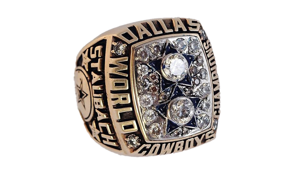 A gold Dallas Cowboys Super Bowl ring with the name Staubach inscribed on the side. The ring features star designs and is encrusted with diamonds, with the words "World Champions" prominently displayed.