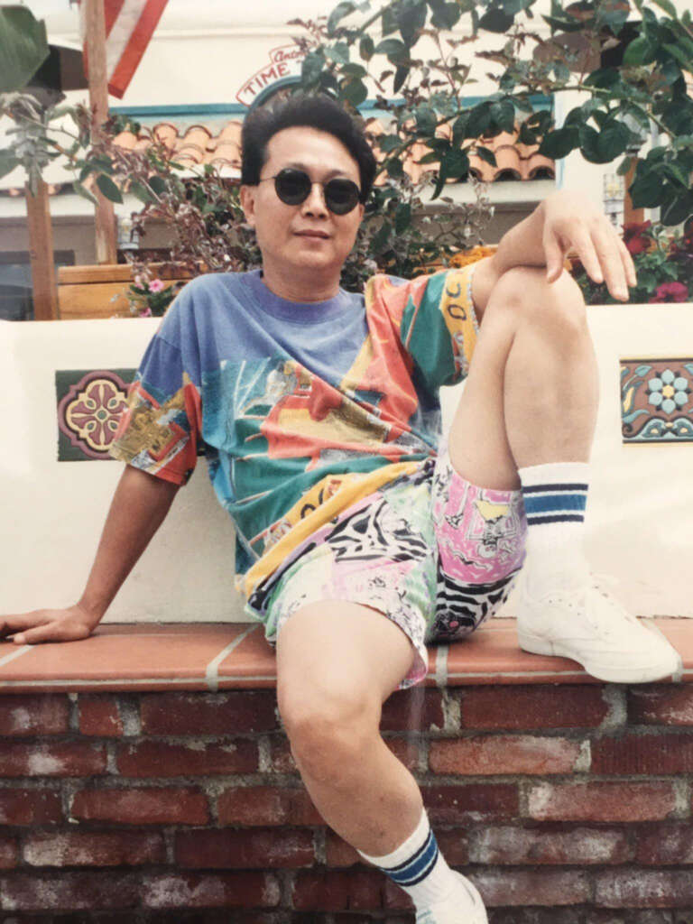 A person in sunglasses and a colorful outfit sits casually on a brick wall. The outfit features a multicolored shirt and bright patterned shorts. White sneakers and striped socks complete the look. There are plants and a tiled wall in the background.