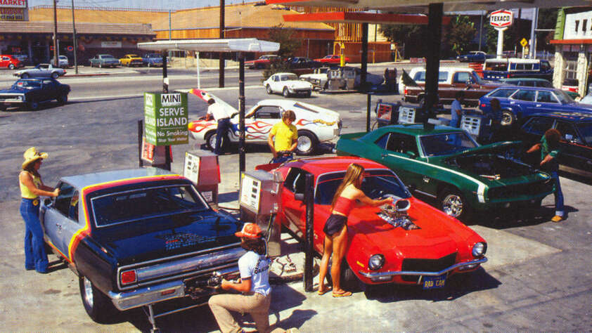 A vintage scene at a bustling gas station with multiple customers fueling their classic cars. The vehicles have distinct colors and hood modifications. Nearby, a gas station sign and retro buildings add to the nostalgic atmosphere.