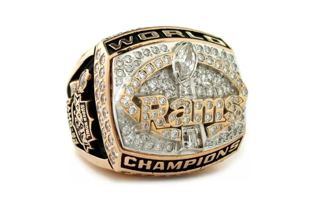 A gold and silver championship ring with "World Champions" inscribed. It features a logo with "Rams" in the center surrounded by diamonds. The sides have detailed engravings, including a stadium image.