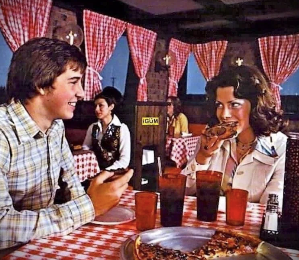 A retro scene showing two people at a pizzeria with red checkered tablecloths and curtains. They are smiling and eating pizza. Glasses of soda are on the table, and the restaurant has a cozy, vintage ambiance.