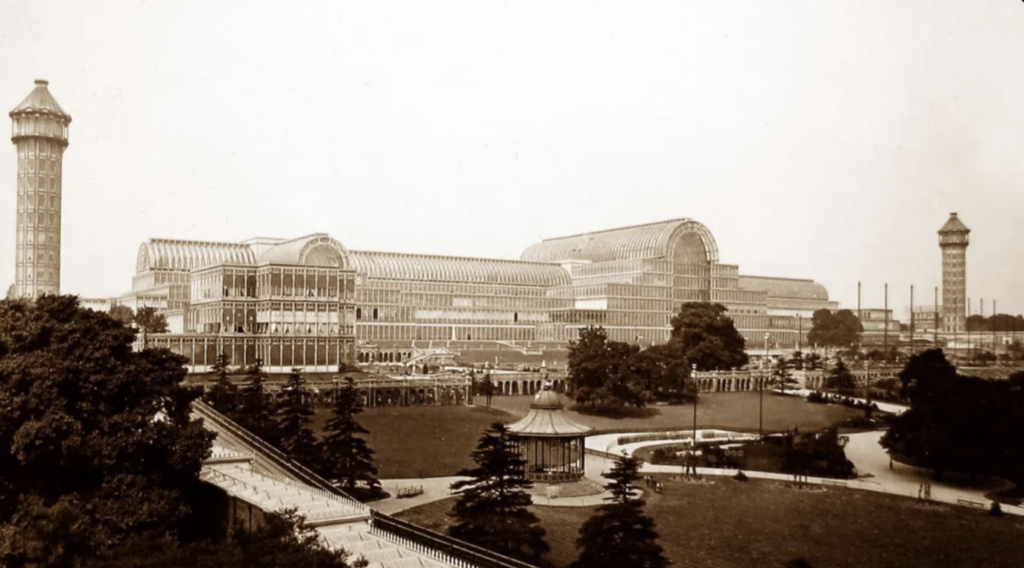 Sepia-toned photo of a large, glass-paneled building with ornate architecture, surrounded by trees and a park. There are three prominent domes and a tower on each side. Pathways lead through the manicured gardens.