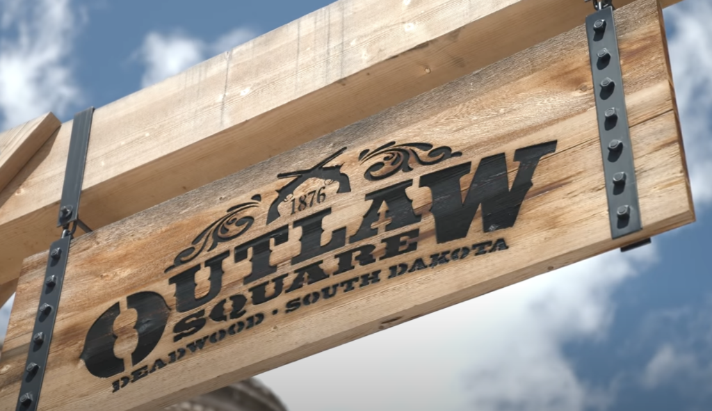 Wooden sign for "Outlaw Square" in Deadwood, South Dakota, featuring a silhouette of a cowboy on horseback above the text. The sign is mounted on a wooden beam with metal brackets, set against a cloudy sky background.