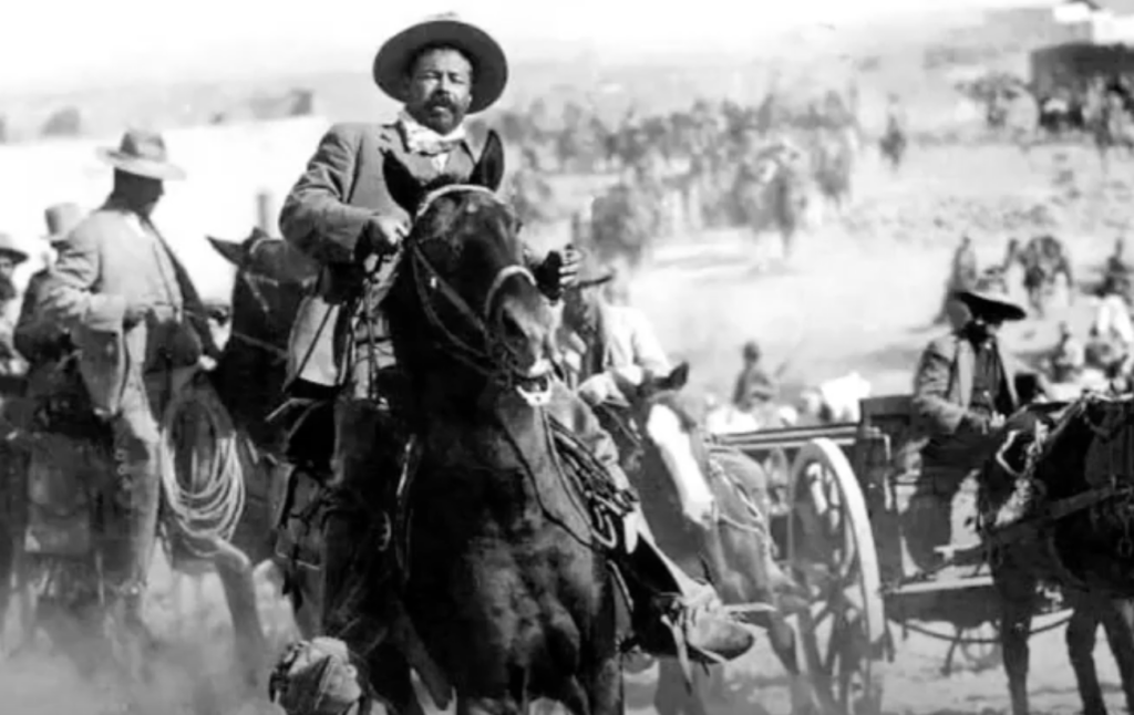 A man wearing a wide-brimmed hat sits confidently on a dark horse in the foreground. Behind him, a group of people on horseback can be seen, with some dust in the air, creating a dynamic scene typical of a historical or western setting.