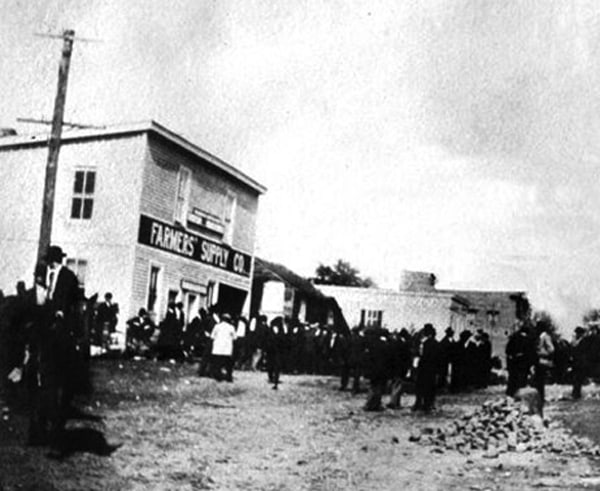 Black and white photo of a large crowd gathered outside "Farmers Supply Co." on a dirt road. People are wearing early 20th-century clothing. A wooden building is visible, with power lines and rocky ground around. Sky appears cloudy.