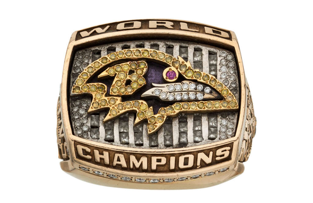 A championship ring with the Baltimore Ravens logo encrusted with yellow and clear stones. "WORLD" is inscribed at the top and "CHAMPIONS" at the bottom, surrounding a textured metallic background.