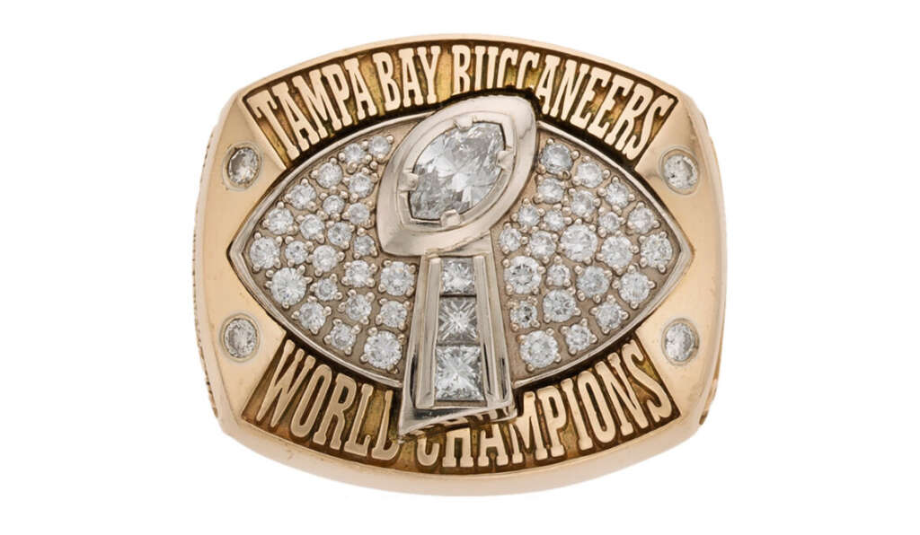 A gold championship ring with the text "Tampa Bay Buccaneers World Champions." It features a large, football-shaped cluster of diamonds and a trophy-like structure in the center, symbolizing victory.