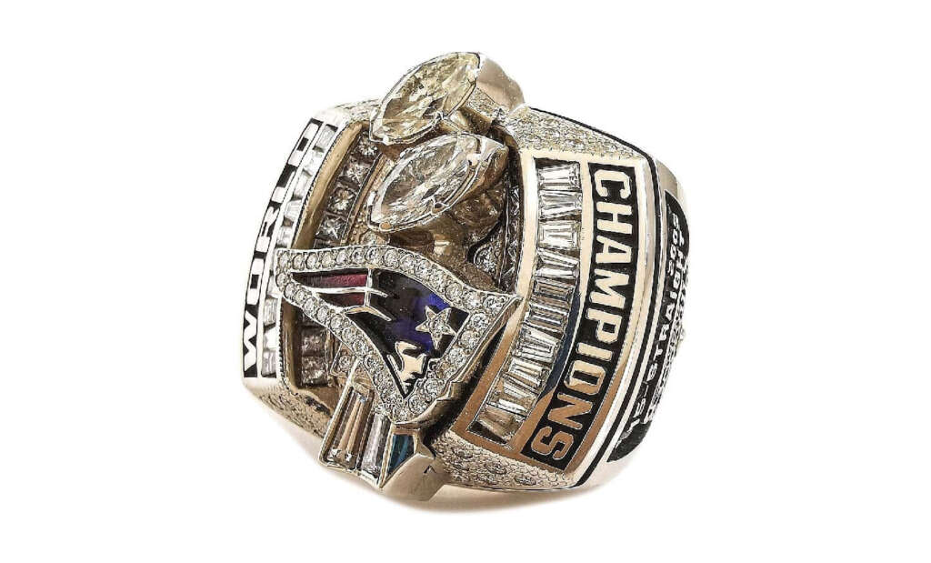 A large, ornate championship ring featuring multiple diamonds and the stylized logo of a patriot's head. The ring has "Champions" inscribed and intricate detailing around the band.