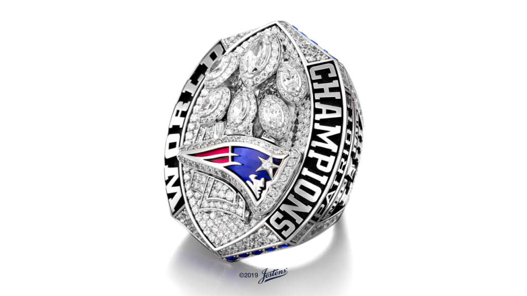 A detailed sports championship ring featuring a blue and red logo at the center surrounded by diamond shapes. The words "WORLD CHAMPIONS" are engraved around the edges. The ring is silver with intricate designs.