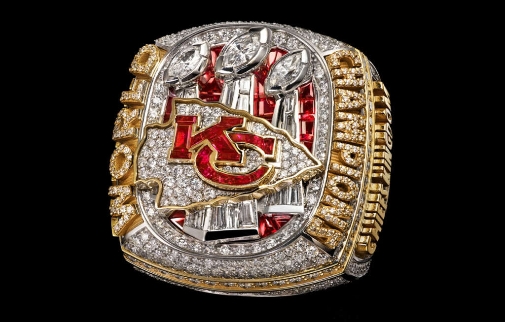 A championship ring encrusted with diamonds and rubies featuring a prominent red "KC" logo in the center. The words "WORLD" and "CHAMPIONS" are engraved on the sides. The background is black, highlighting the ring's sparkle.