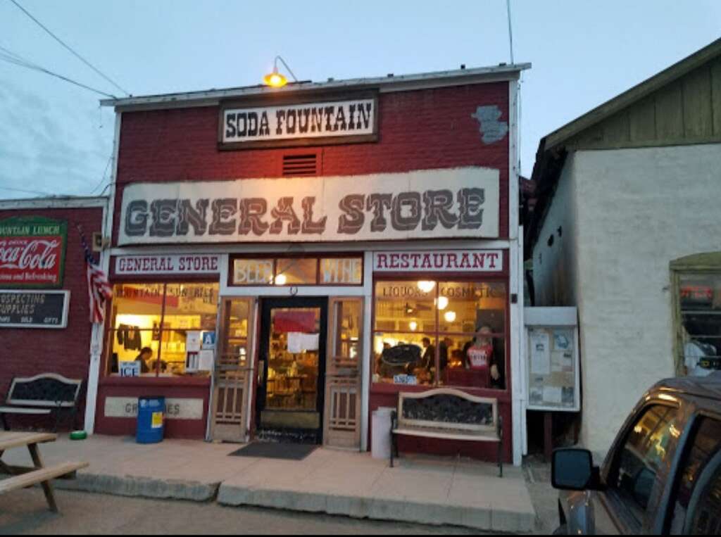 Vintage general store with a soda fountain, displaying signs for a restaurant and beer/wine. The facade is rustic, with a Coca-Cola sign and American flag. A car and picnic table are visible in the foreground.